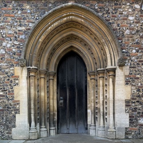 A pointed Gothic-style black wooden chapel door set within a deeply recessed stone archway of a flint and brick wall.