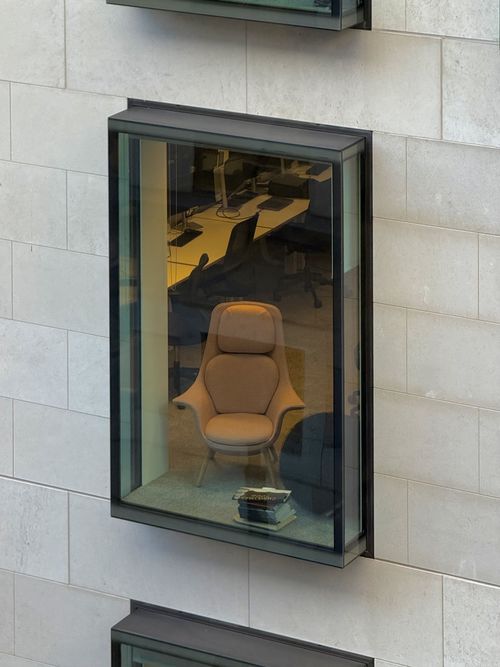 A glass box window in a light stone building reveals a single cushioned armchair facing outwards towards the view. There’s a small stack of books next to the chair, while an office workspace with desks, monitors, and rolling chairs is visible in the background. To edges of the box windows above and below are just visible.